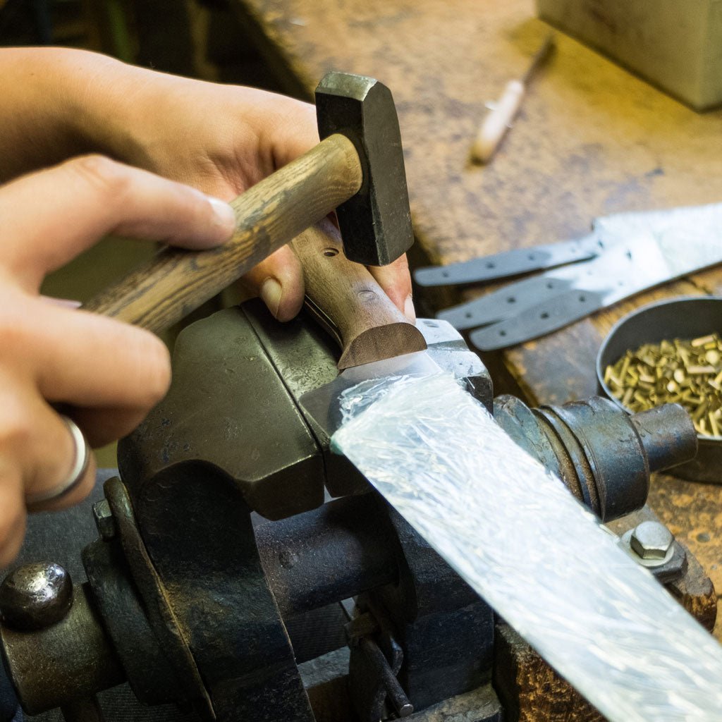 Hand using a hammer on a wooden knife handle in a workshop setting
