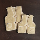 Two cream-colored children's woolen vests on a dark background. 