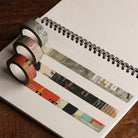 Three rolls of decorative washi tape with vintage-style collage patterns and text, arranged on an open spiral notebook.
