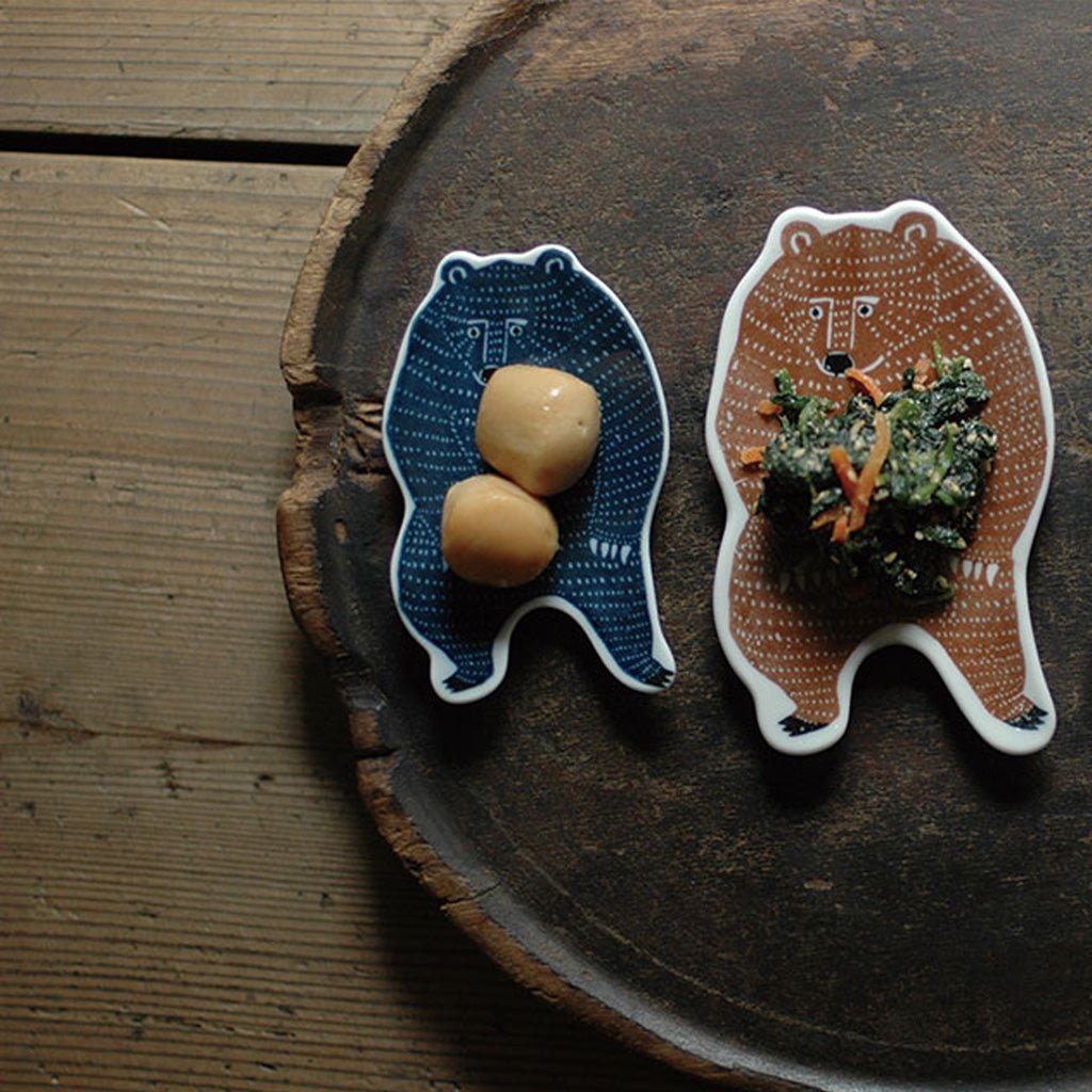 Two bear-shaped ceramic serving dishes on a rustic wooden board, one blue with white dots and one brown with white dots, styled with food on a wooden table.