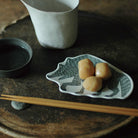 A small white ceramic plate with a dark gray wolf illustration holds three peach-colored pieces of food, set on a rustic dark wooden tray with chopsticks and a white cup in the background.