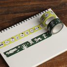 Two rolls of washi tape with cartoon animal patterns, one in light yellow-green and one in dark green, displayed on an open spiral notebook over a wooden surface.