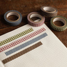 Four rolls of washi tape with gingham and polka dot patterns in muted purple gray, dusty blue, green, and brown, arranged on a wooden surface next to a grid notebook with strips of the tape applied.