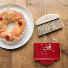 Baked pastry with orange slices and red berries on a plate next to a table brush and red dustpan on a wooden surface.