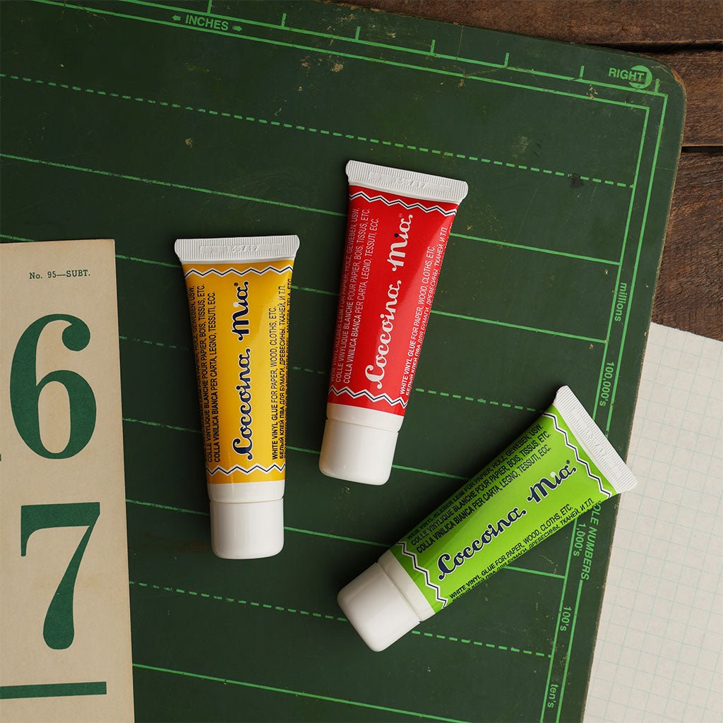 Three colored tubes of Coccoina Mia White Vinyl Glue Adhesive on a green chalkboard