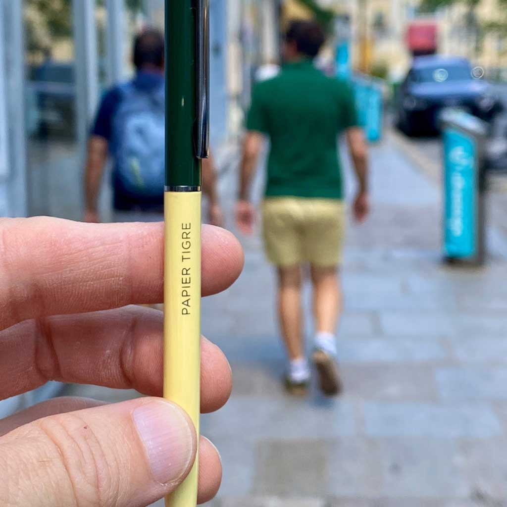 Hand holding a yellow pen with 'PAPIER TIGRE' branding against a blurred street background