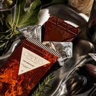 A nightcap chocolate bar open next to metal barware and herbs.