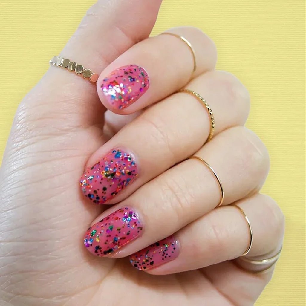 Hand wearing pink nail polish with rainbow confetti cake glitter top coat against yellow background
