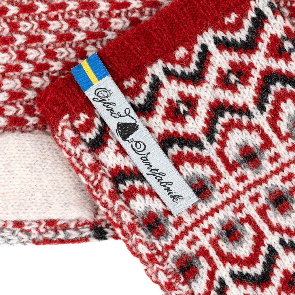 Close-up of a knitted textile with a red, cream, black, and gray Fair Isle pattern, featuring a white fabric label with the brand name 'Ojbro Vantfabrik' and a small Swedish flag.