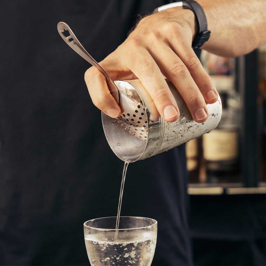 Person pouring liquid from a metal shaker through julep strainer into a glass with a blurred background