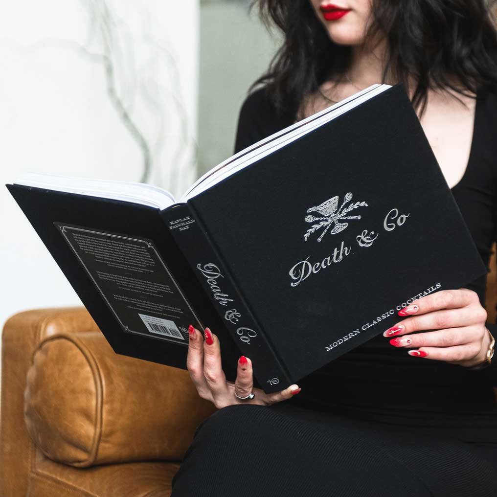 Person holding a black book titled 'Death & Co' with a logo on a blurred background.