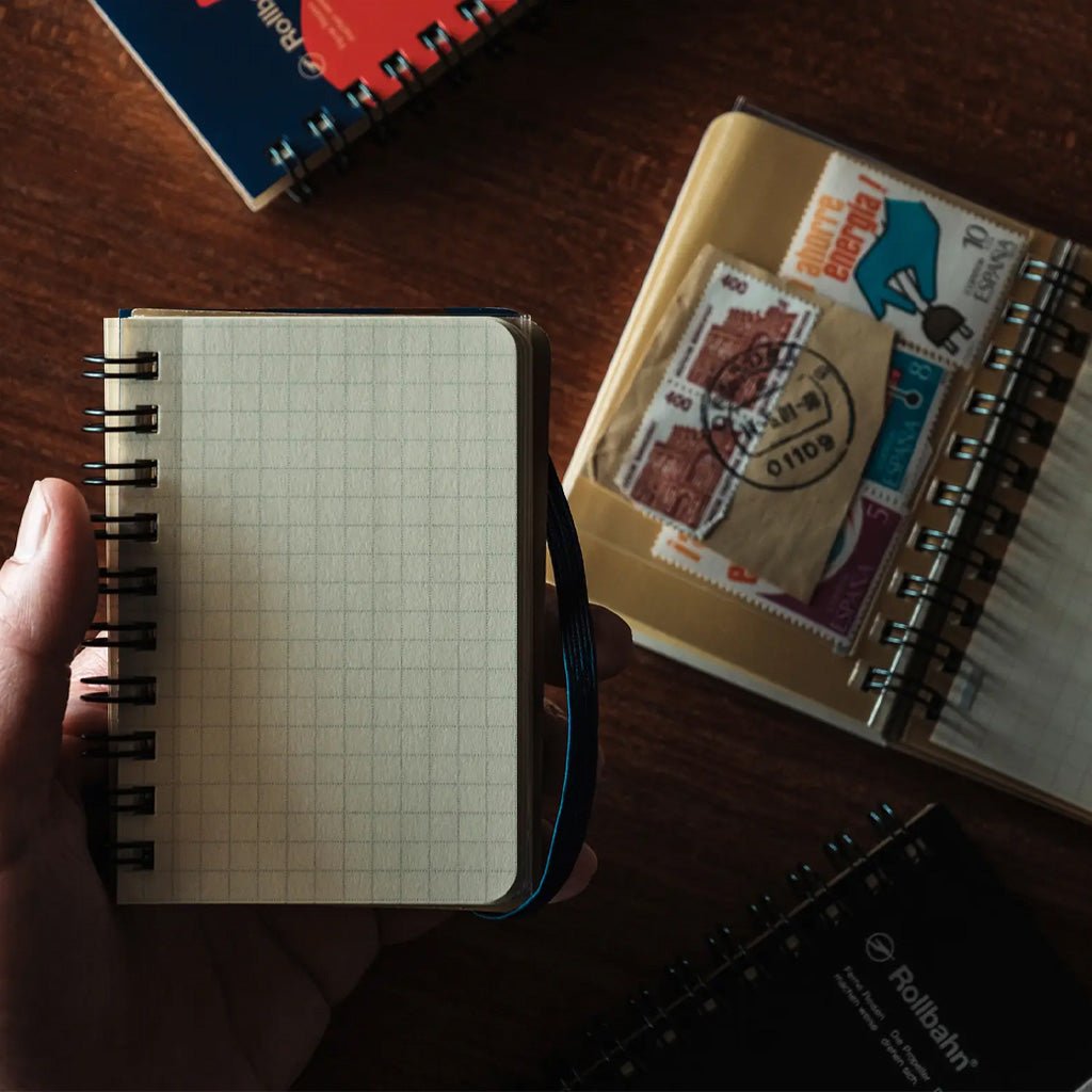 A hand holds a small open grid notebook with black spiral binding on a dark wooden table. Nearby are other notebooks and a clear pocket organizer containing postage stamps.