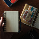 A hand holds a small open grid notebook with black spiral binding on a dark wooden table. Nearby are other notebooks and a clear pocket organizer containing postage stamps.