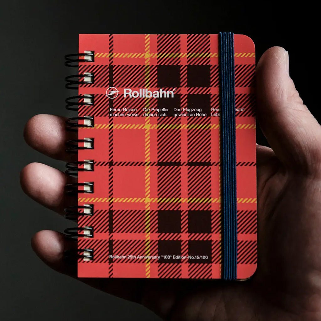 A hand holding a small Rollbahn spiral notebook with a red and black tartan plaid cover and a dark blue elastic closure band, against a dark background.