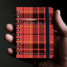 A hand holding a small Rollbahn spiral notebook with a red and black tartan plaid cover and a dark blue elastic closure band, against a dark background.