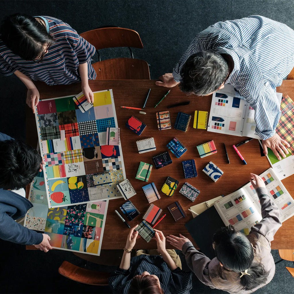 Overhead view of several people gathered around a wooden table, examining colorful designs, patterned paper samples, notebooks, and art books in a creative studio setting.