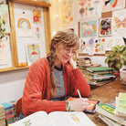 Woman sitting at desk illustrating on a tablet, surrounded by artwork.