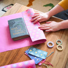 Hand wrapping book with wrapping paper next to colorful supplies.