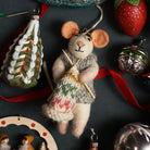 Mouse ornament holding a knitting project with Christmas decorations on a dark background.
