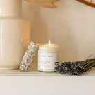Fern & Moss candle with a lit wick, surrounded by dried lavender and sage on a neutral background