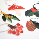 Close up of set of Christmas-themed gift tags including a mushroom, holly, pine cone, and berries on a white background.
