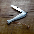 A folding pocket knife with a fish-shaped silver metal handle and a silver blade, resting on a rustic wooden surface with a brown leather lanyard.