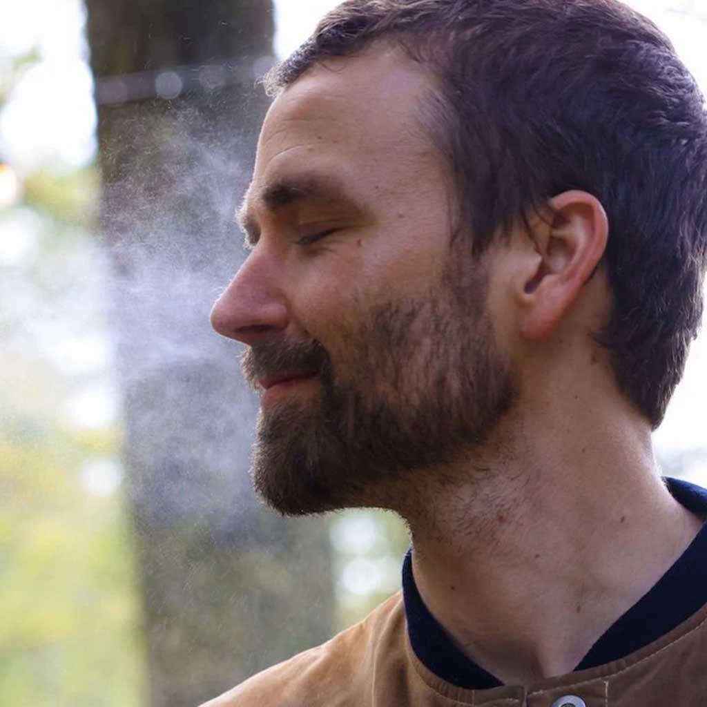 Man with a beard and short hair standing outdoors spraying facial toner with a blurred natural background