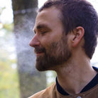 Man with a beard and short hair standing outdoors spraying facial toner with a blurred natural background