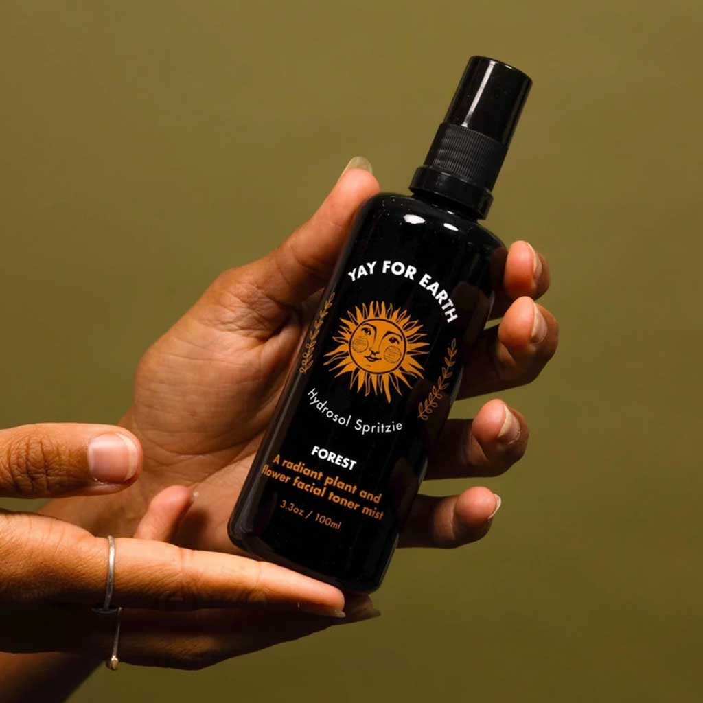 Hand holding a black bottle of hydrosol toner labeled 'Yay for Earth' against a green background
