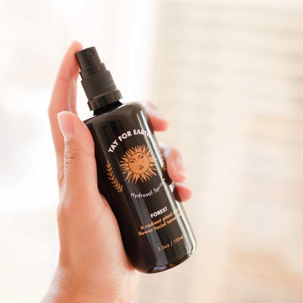 Hand holding a black spray bottle of hydrosol toner labeled 'Yay for Earth' with a sun design on a blurred background