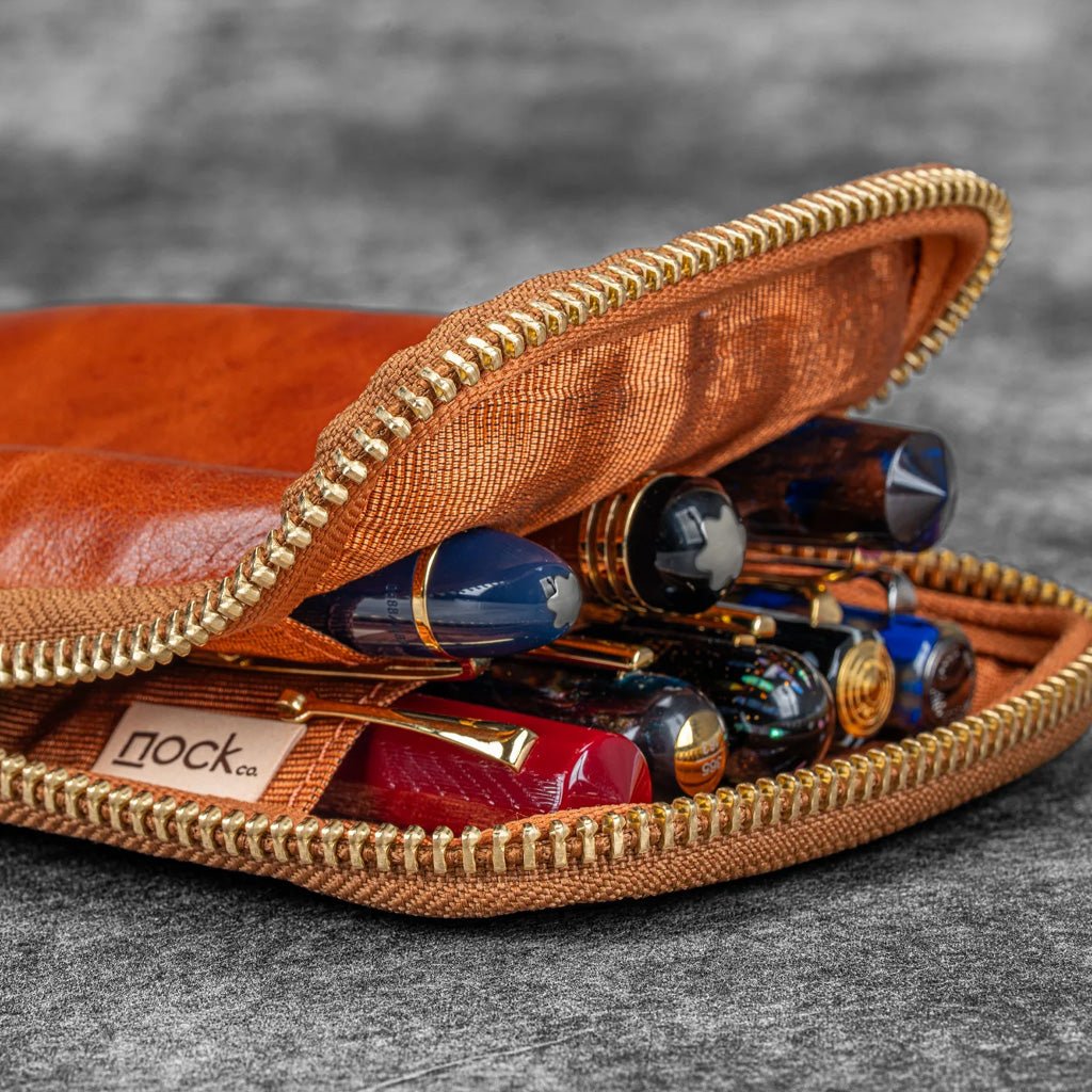 Brown leather case with gold zipper containing various pens on a gray background