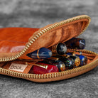 Brown leather case with gold zipper containing various pens on a gray background