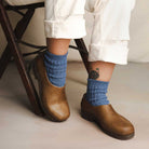 Person wearing blue socks and brown hemp clog shoes with white pants, sitting on a wooden chair.