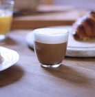 Cappuccino in a glass on a wooden table with a croissant and orange juice in the background.