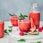 Glass of watermelon juice with a bottle and additional glasses on a marble surface.