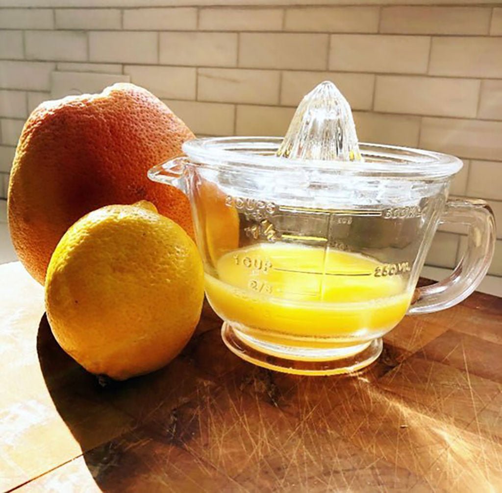 Clear glass juicer with orange juice next to a grapefruit and lemon on a wooden surface.