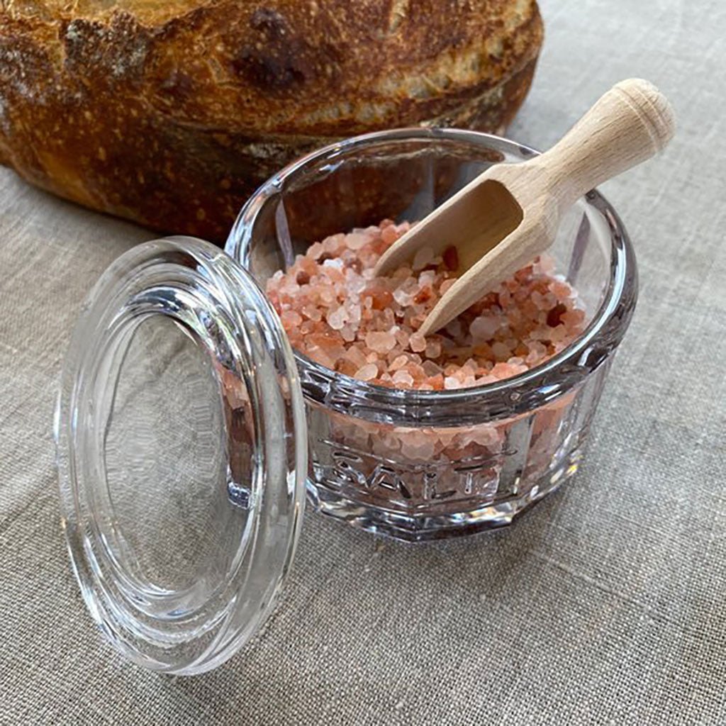 Glass jar of pink himalayan salt with a wooden scoop on a tablecloth, set near a loaf of bread.