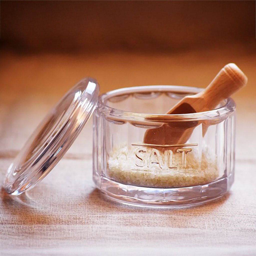 A clear glass salt container with a matching glass lid, filled with coarse salt and topped with a small wooden scoop.