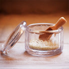 A clear glass salt container with a matching glass lid, filled with coarse salt and topped with a small wooden scoop.