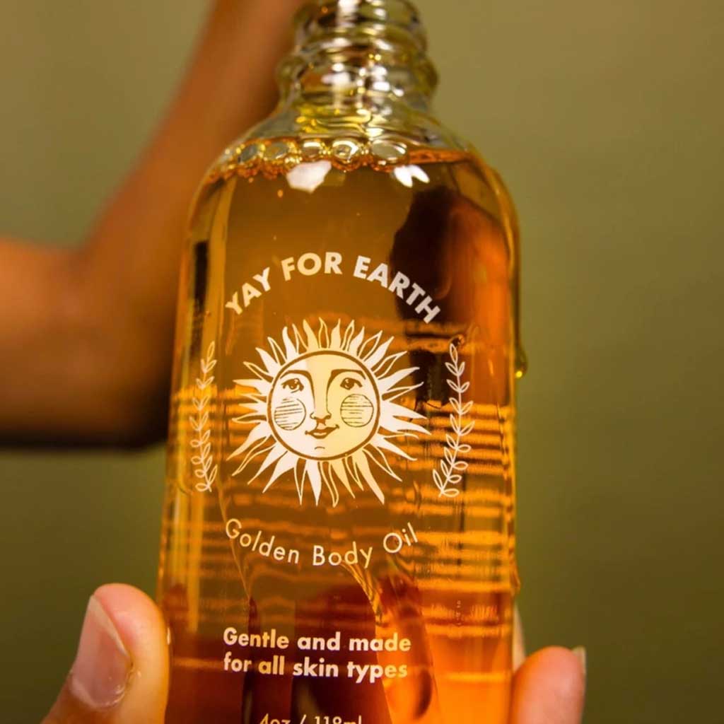 Bottle of 'Yay for Earth' Golden Body Oil held by a person against a green background