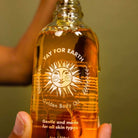 Bottle of 'Yay for Earth' Golden Body Oil held by a person against a green background
