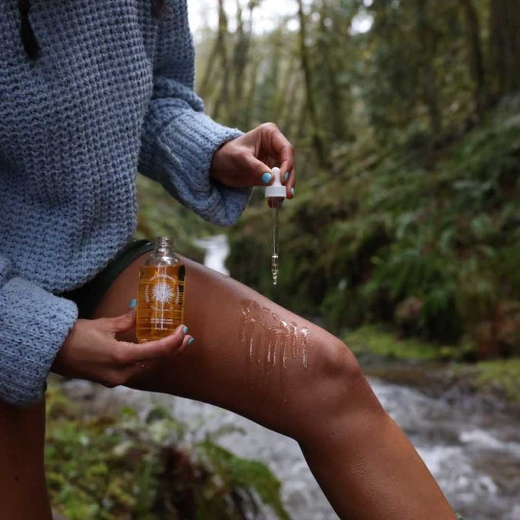 Person applying a natural body oil product outdoors with a forest background