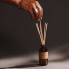Hand with rings holding reed diffuser sticks over a bottle on a dark background