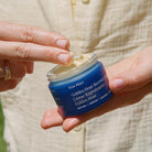 A close-up of a person holding a blue jar of Ursa Major Golden Hour Recovery Cream, with their fingers dipping into the cream.