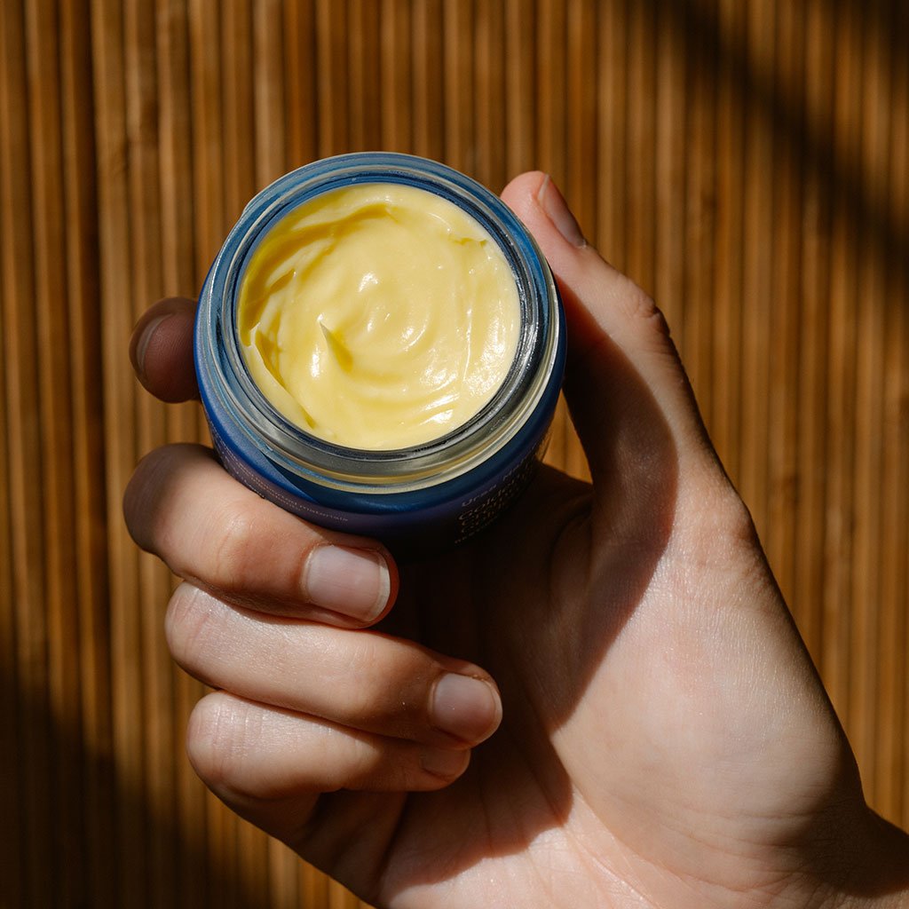 A hand holding an open jar of pale yellow cream.