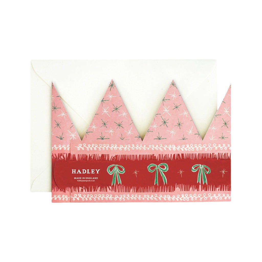 A pink and red crown-shaped greeting card with pointed peaks, decorated with small white stars, green bows, and white borders. A white envelope is partially visible behind the card.