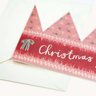 A close-up of a Christmas greeting card with a crown design, featuring the word 'Christmas' in white script on a red band, with a green bow illustration and delicate white decorative borders. The card is partially layered over a white envelope on a clean white background.