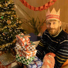 A man wearing a paper crown that says 'Christmas Fairy' sits next to a decorated Christmas tree and a stack of wrapped gifts, holding a plaid Christmas stocking.