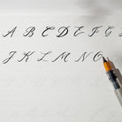 Black calligraphy ink shown with a sample alphabet on white paper, with part of a black and orange calligraphy pen visible in the lower right corner.