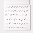 A square white book page showing a cursive alphabet in black ink, with uppercase letters across the top rows and lowercase letters across the bottom rows, photographed against a light neutral background.